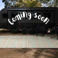 A black caravan parked outdoors on a gravel surface with a green camping recycled mat  two black camping chairs, and a small side table. Large white text reading 'Coming soon' is overlaid across the trailer.