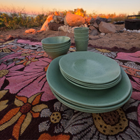 A set of speckled sage green plates and bowls arranged on a colorful Sunblossom woven trqvel rug outdoors at sunset, with rocks and a campfire in the background. asymmetrical dinnerware