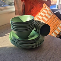 A set of speckled matte green rPET tableware, including bowls, plates, and cups, stacked on a gray surface in natural sunlight with a cozy home background featuring patterned pillows in a caravan. asymmetrical dinnerware