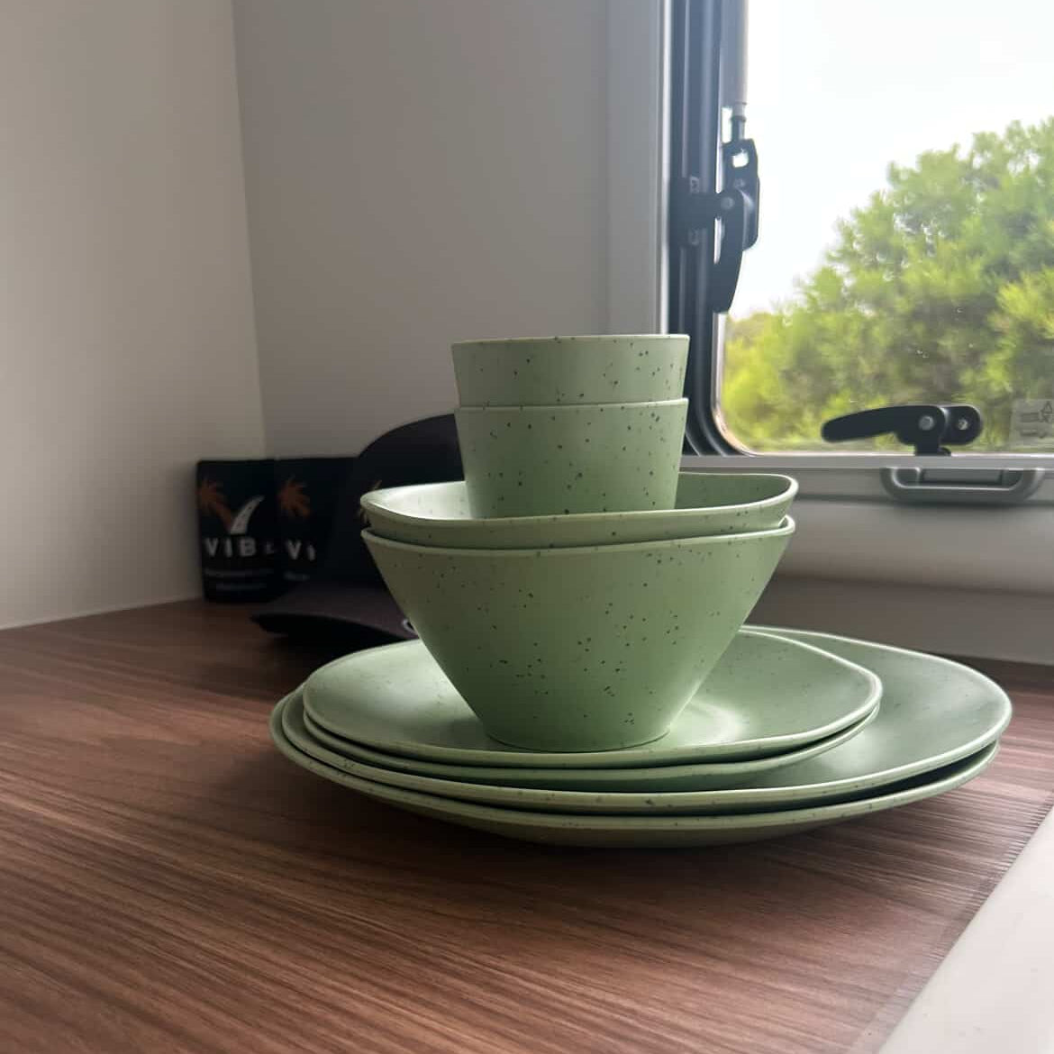 Stack of Speckled green rPET bowls and plates on a wooden caravan surface with a window in the background. asymmetrical dinnerware