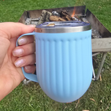Hand holding a blue mug with a silver lid over a campfire