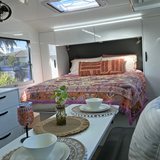 Speckled Coconut white rPET caravan dinnerware styled on a caravan table with matching wine tumbler, indoor plant and Dahlia woven throw rug on the bed, showcasing a practical and stylish vanlife setup. asymmetrical dinnerware