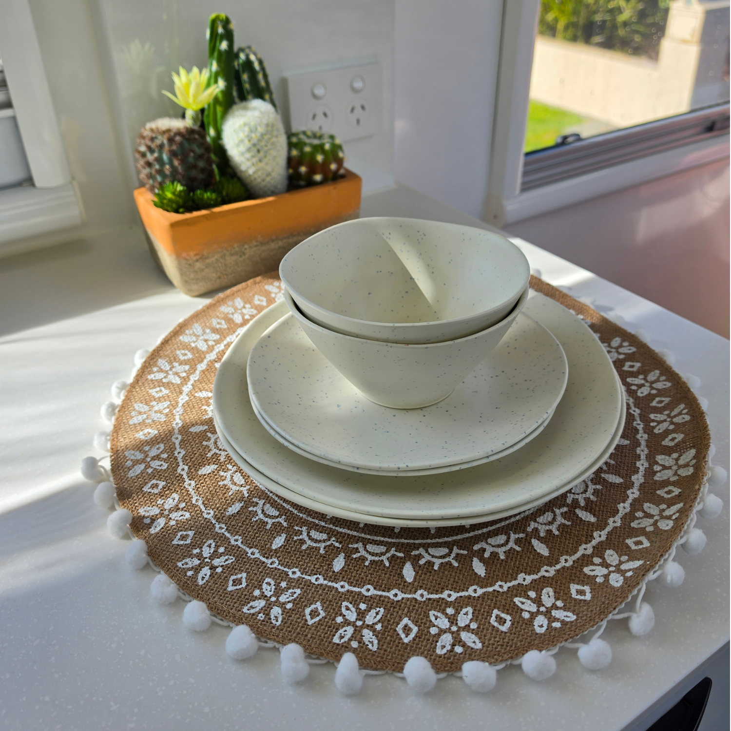 Speckled Coconut white rPET caravan dinnerware set including dinner plate, side plate and two bowls, styled on a woven boho placemat beside a cactus planter in natural sunlight. asymmetrical dinnerware