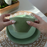 Hands gently flexing the Vanaway Speckled Matcha rPET bowl to demonstrate its lightweight, durable and shatter-resistant design, with matching dinnerware underneath. asymmetrical dinnerware