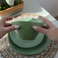 Hands gently flexing the Vanaway Speckled Matcha rPET bowl to demonstrate its lightweight, durable and shatter-resistant design, with matching dinnerware underneath. asymmetrical dinnerware