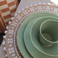 Close-up of Vanaway Speckled Matcha rPET caravan dinnerware, showing the speckled sage green plate, side plate, bowl and tumbler stacked on a decorative woven placemat. asymmetrical dinnerware