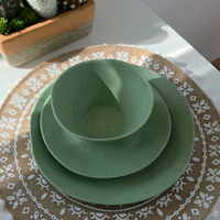 Top-down view of Vanaway Speckled Matcha rPET caravan dinnerware featuring a speckled sage green dinner plate, side plate and bowl stacked on a woven placemat with natural sunlight. asymmetrical dinnerware