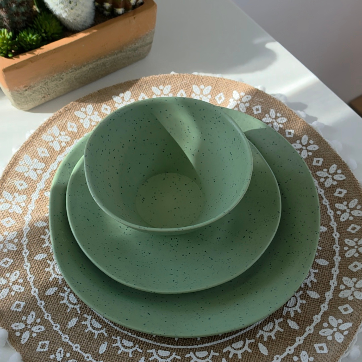 Top-down view of Vanaway Speckled Matcha rPET caravan dinnerware featuring a speckled sage green dinner plate, side plate and bowl stacked on a woven placemat with natural sunlight. asymmetrical dinnerware