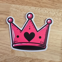 A die-cut sticker of a pink crown with a black heart in the center, placed on a light wood grain surface.