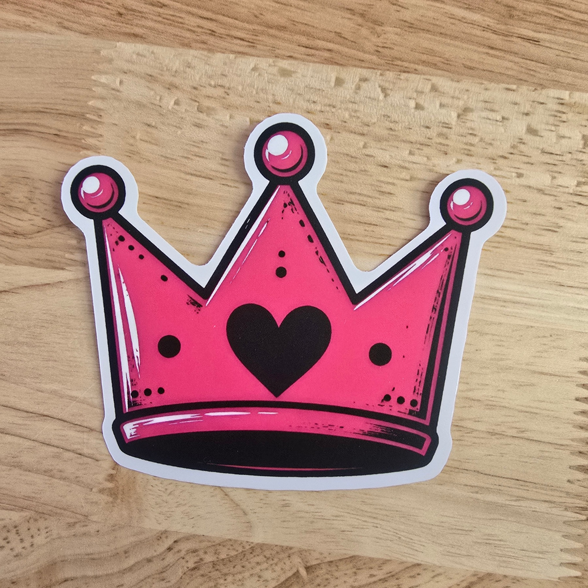 A die-cut sticker of a pink crown with a black heart in the center, placed on a light wood grain surface.