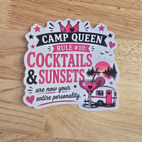A die-cut sticker with a whimsical graphic design reading 'CAMP QUEEN RULE #10: COCKTAILS & SUNSETS are now your entire personality.' The sticker features pink and black text, a crown, a cocktail glass, a sunset, and a retro trailer, placed on a light wood surface.