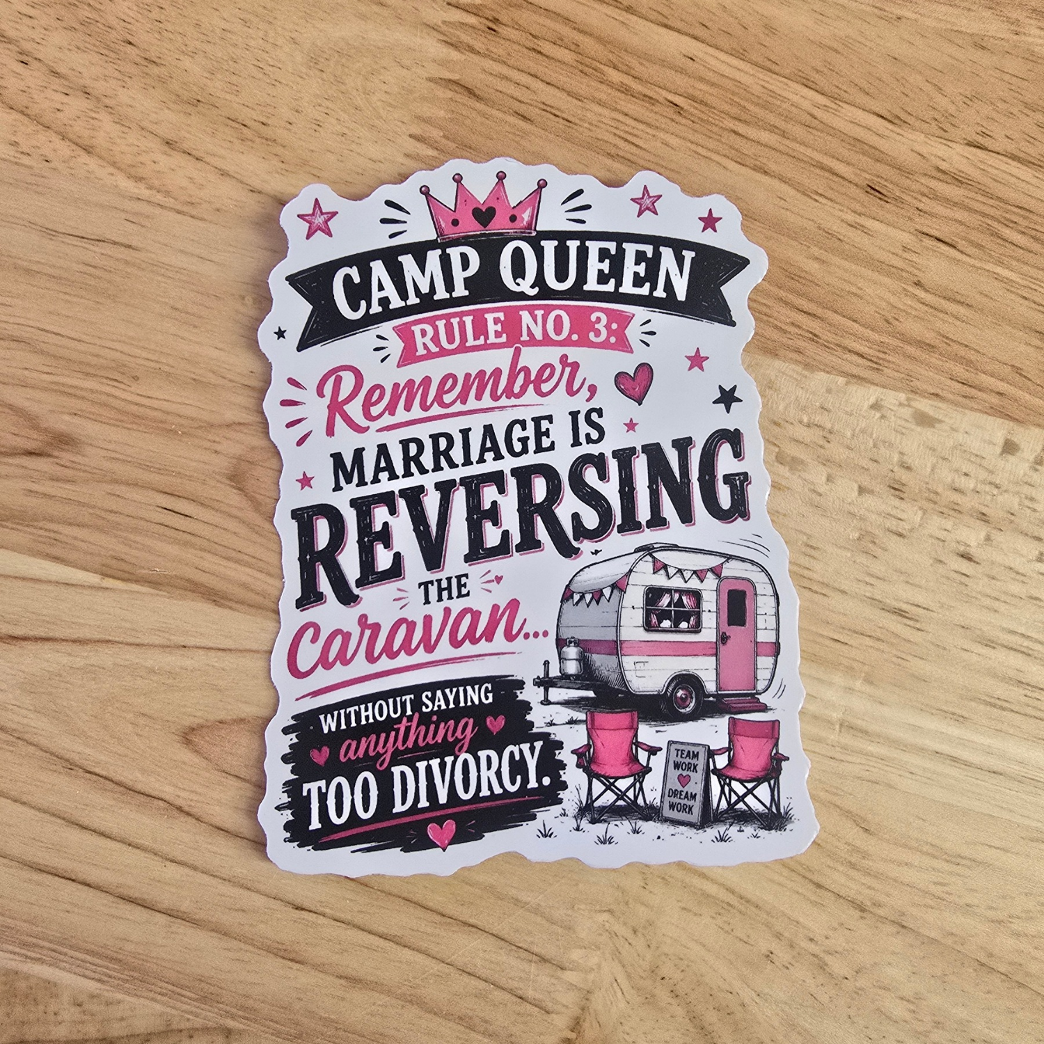 A die-cut camp queen sticker with a humorous quote about camp queens and divorce, featuring a retro caravan illustration, placed on a light wood surface.