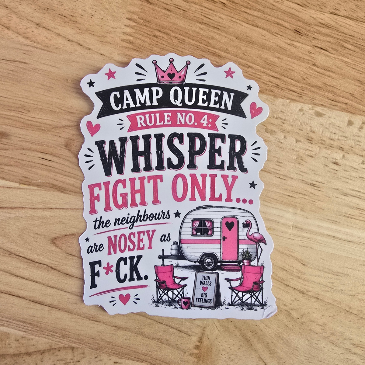 A die-cut sticker with a humorous camp queen slogan, displayed on a light wood grain surface. The sticker features a white background with pink and black text reading 'CAMP QUEEN RULE NO 4
