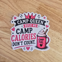 A die-cut sticker with the text 'CAMP QUEEN RULE #6: CAMP CALORIES DON'T COUNT.' and an illustration of a pink camp mug with whipped cream, placed on a light wood surface.