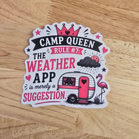A die-cut sticker with a whimsical camping theme, featuring the text 'CAMP QUEEN THE WEATHER APP is merely a SUGGESTION' in black and pink lettering, surrounded by illustrations of a pink trailer, a pink flamingo, and a smiling sun, displayed on a light wood background.