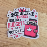 A die-cut sticker featuring a cartoon pink wallet with a crown and the text 'CAMP QUEEN RULE #8: HOLIDAY BUDGETS are FICTIONAL.' placed on a light wood surface.