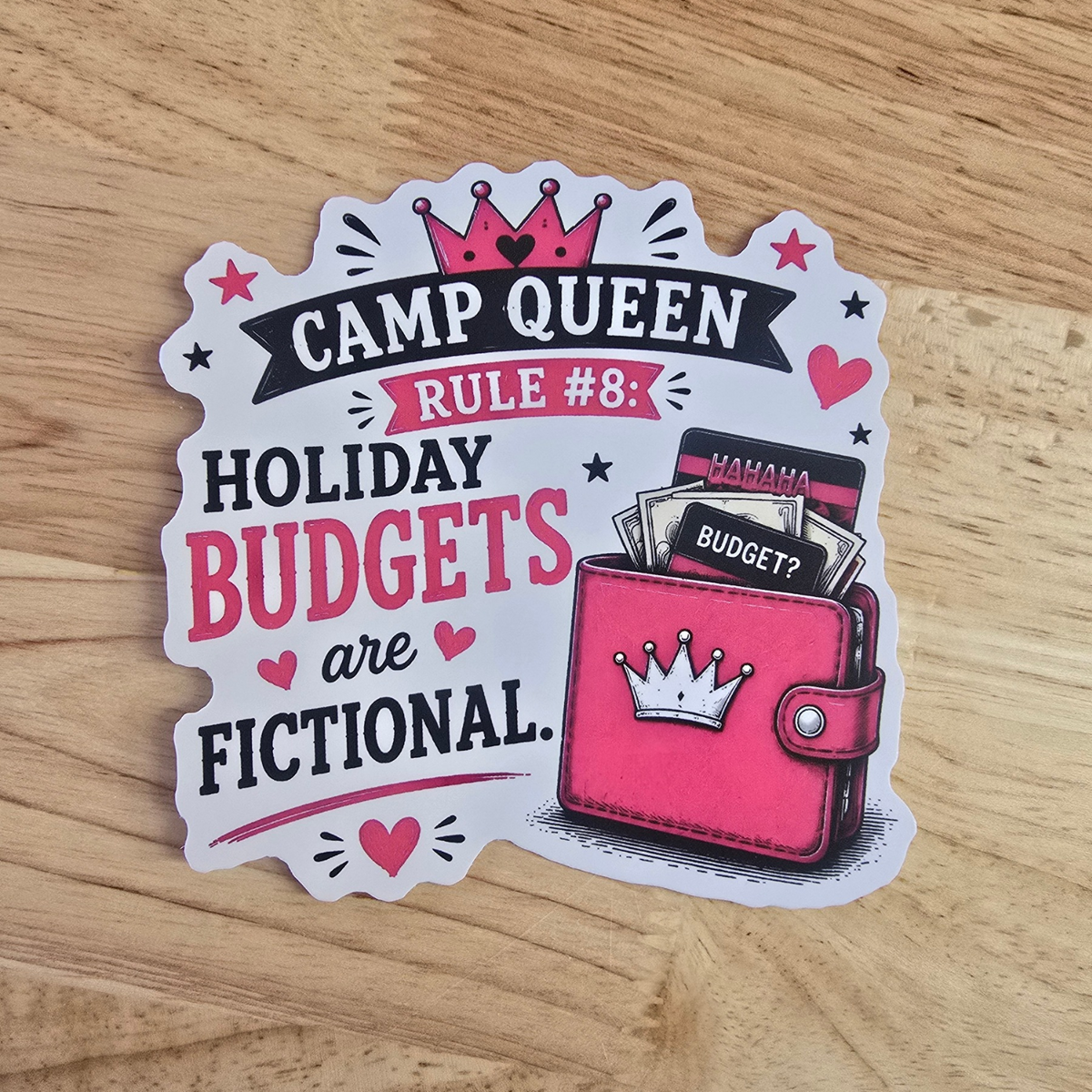 A die-cut sticker featuring a cartoon pink wallet with a crown and the text 'CAMP QUEEN RULE #8: HOLIDAY BUDGETS are FICTIONAL.' placed on a light wood surface.