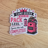 A die-cut sticker with a 'Camp Queen Rule #9' design, featuring pink and black text and an illustration of a pink suitcase, placed on a light wood grain surface.
