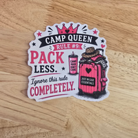 A die-cut sticker with a 'Camp Queen Rule #9' design, featuring pink and black text and an illustration of a pink suitcase, placed on a light wood grain surface.