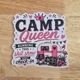 A die-cut sticker with a 'Camp Queen' theme, featuring pink and black text, a retro camper trailer, a pink flamingo, and stars, displayed on a light wood background.