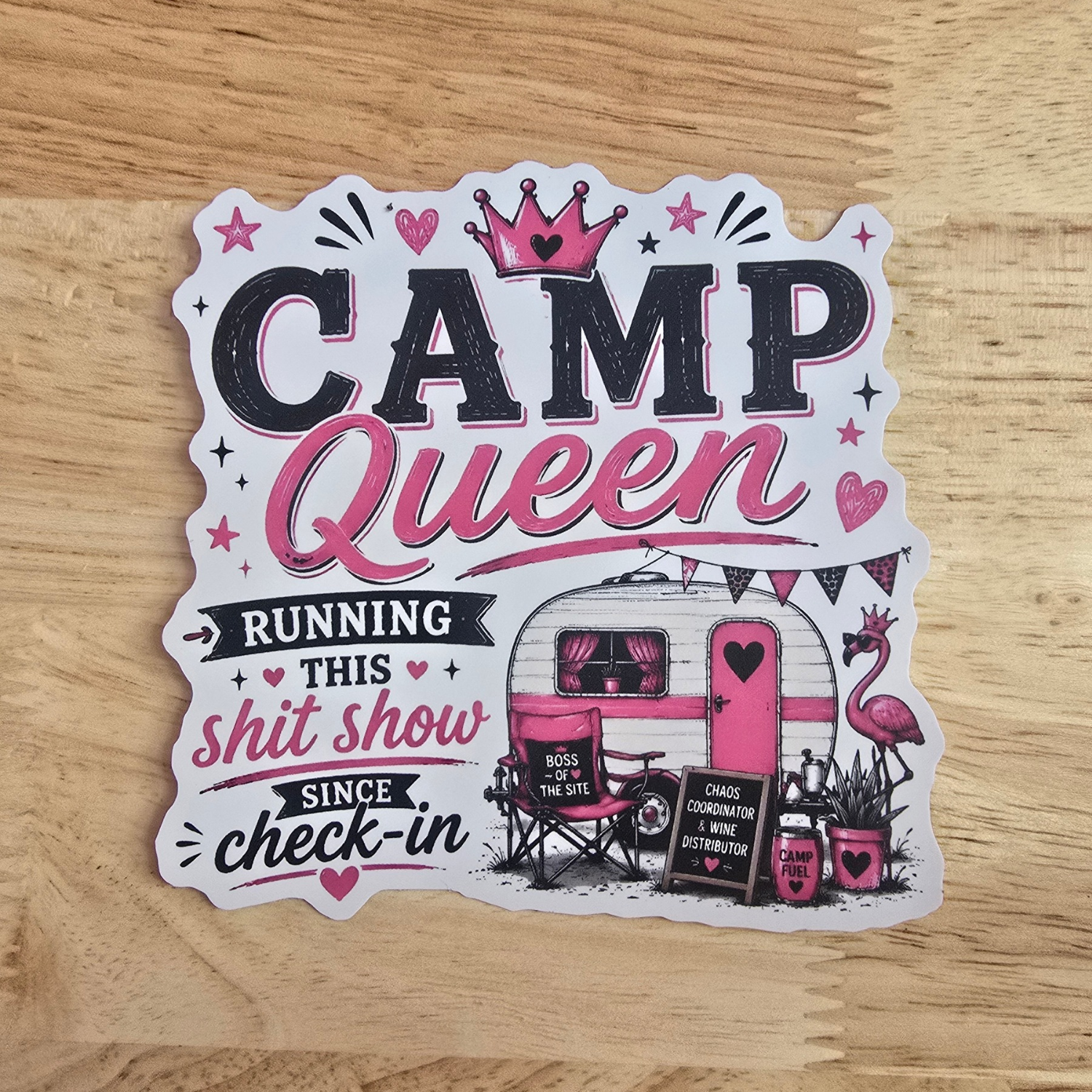 A die-cut sticker with a 'Camp Queen' theme, featuring pink and black text, a retro camper trailer, a pink flamingo, and stars, displayed on a light wood background.