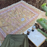 Vanaway Solana woven travel throw rug laid out at a campsite with camping chairs, insulated wine tumblers and a camp table, featuring a mustard, blush and cream palm inspired design.