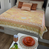 Vanaway Solana woven travel throw rug styled across a campervan bed with matching dinnerware and insulated wine tumbler, featuring a mustard, blush and cream palm inspired design with decorative fringe.