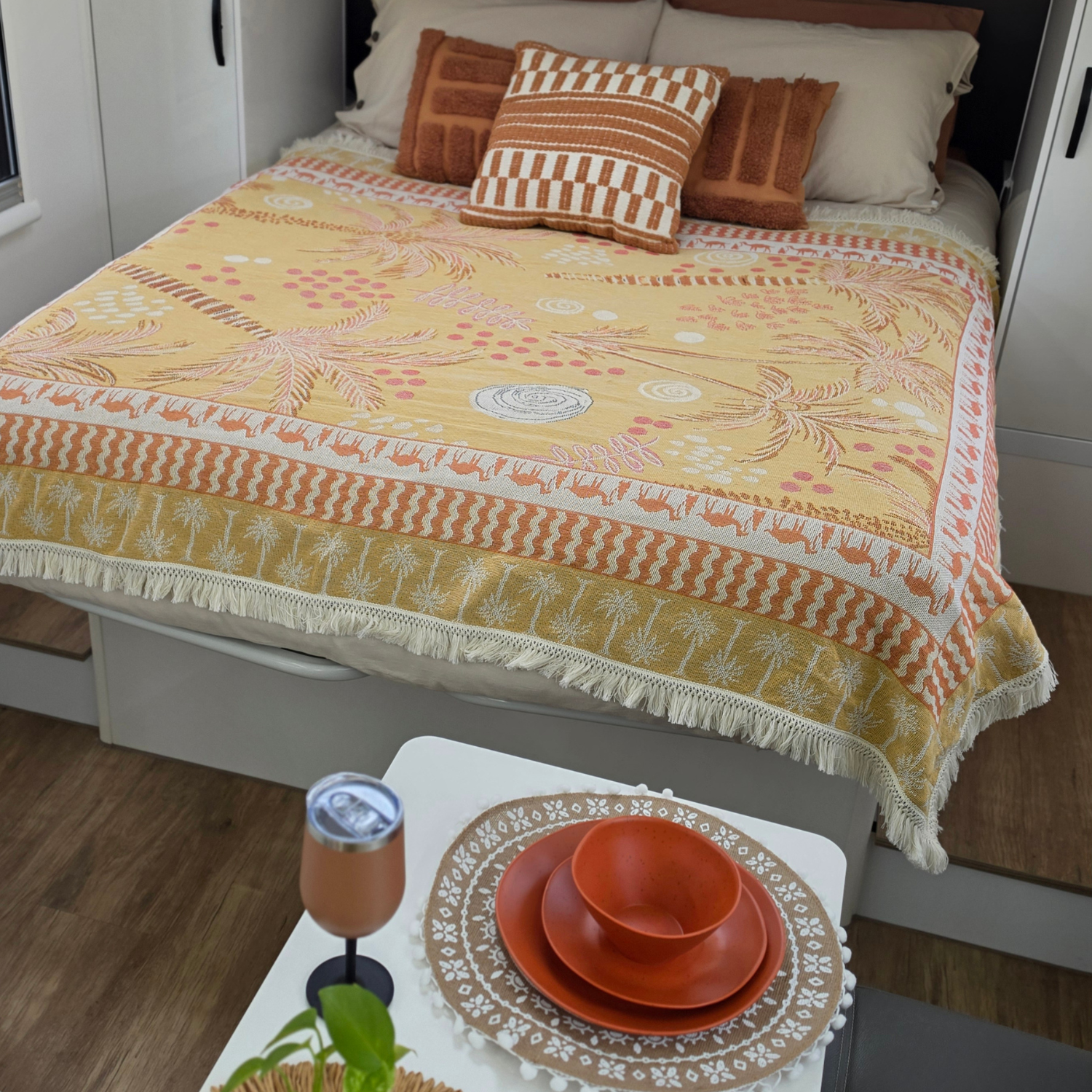 Vanaway Solana woven travel throw rug styled across a campervan bed with matching dinnerware and insulated wine tumbler, featuring a mustard, blush and cream palm inspired design with decorative fringe.