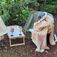 Vanaway Solana woven travel throw rug draped over a camping chair beside a camp table with insulated wine tumblers, showcasing its reversible design, soft woven cotton texture and decorative fringe.