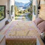 Vanaway Solana woven travel throw rug styled across a campervan bed with a warm palm inspired design in mustard, blush and cream overlooking a mountain landscape.
