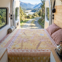 Vanaway Solana woven travel throw rug styled across a campervan bed with a warm palm inspired design in mustard, blush and cream overlooking a mountain landscape.