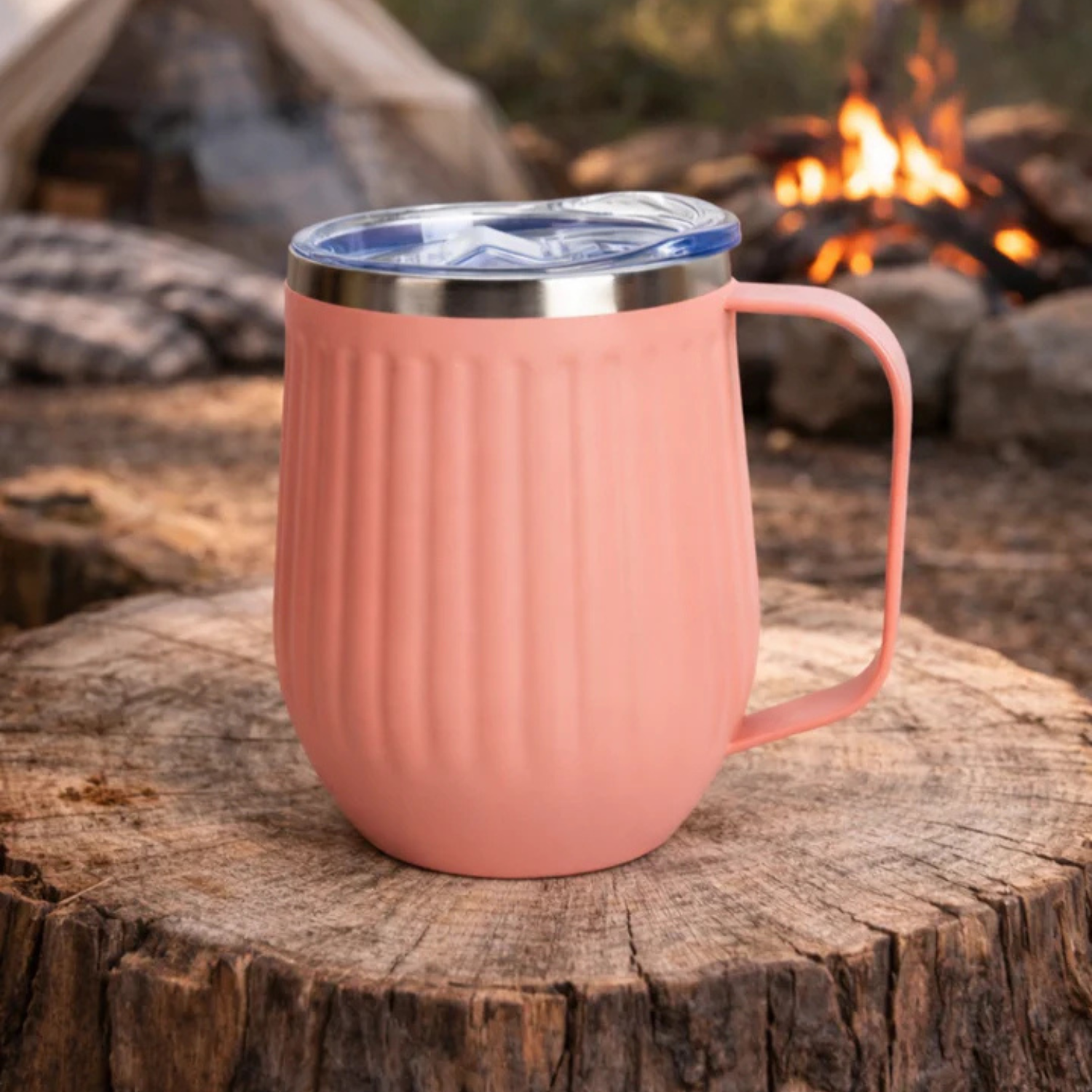 Vanaway Blush ribbed insulated camping mug with a spill resistant lid and stainless steel rim, styled on a tree stump beside a campfire for cosy camping adventures.