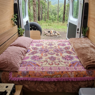 Vanaway Dahlia woven travel throw rug featuring a cream, lilac, lavender and terracotta boho paisley design, styled as a decorative bed throw inside a campervan overlooking a peaceful bush campsite.