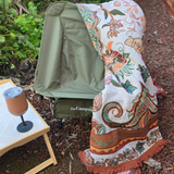 Vanaway Sahara woven travel throw rug featuring a cream, terracotta, sage green and blush boho botanical design, draped over a camping chair beside a camp table for cosy outdoor adventures.