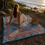Vanaway Santorini woven travel throw rug featuring a soft blue, terracotta and cream boho paisley design, styled by the coast at sunset for caravanning, camping and beach adventures.