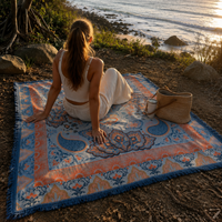 Vanaway Santorini woven travel throw rug featuring a soft blue, terracotta and cream boho paisley design, styled by the coast at sunset for caravanning, camping and beach adventures.