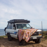 Vanaway Sunblossom woven travel throw rug draped over the bonnet of a 4WD parked beside the beach, showcasing the vibrant floral design in an outdoor coastal setting.