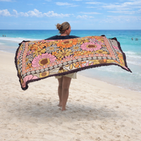 Woman carrying the Vanaway Sunblossom woven travel throw rug along a white sandy beach, showcasing the vibrant floral design by the turquoise ocean.