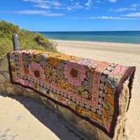 Vanaway Sunblossom woven travel throw rug draped over a beach lookout wall, showcasing the vibrant floral design with the ocean in the background.