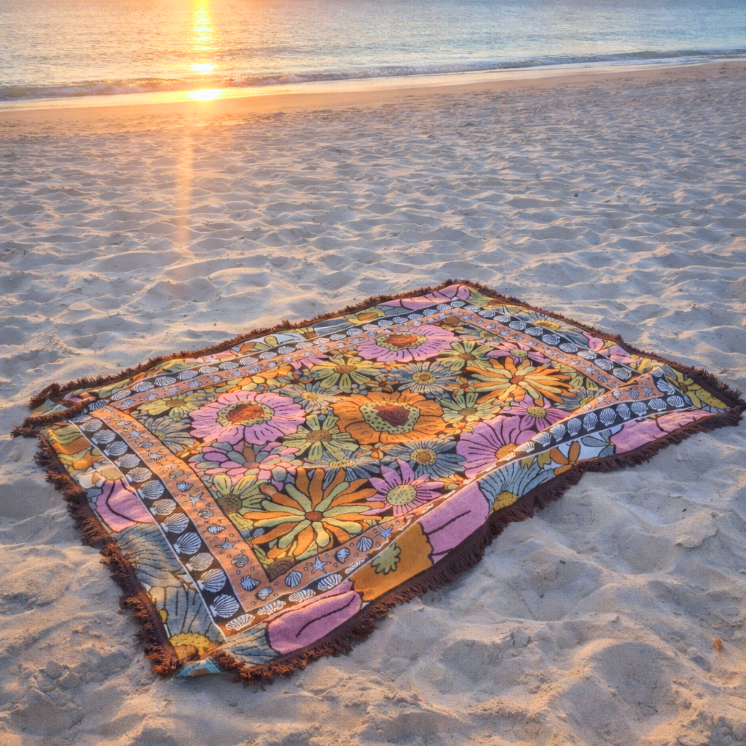 Vanaway Sunblossom woven travel throw rug laid on soft sand at the beach during sunset, showcasing the vibrant floral design by the shoreline.