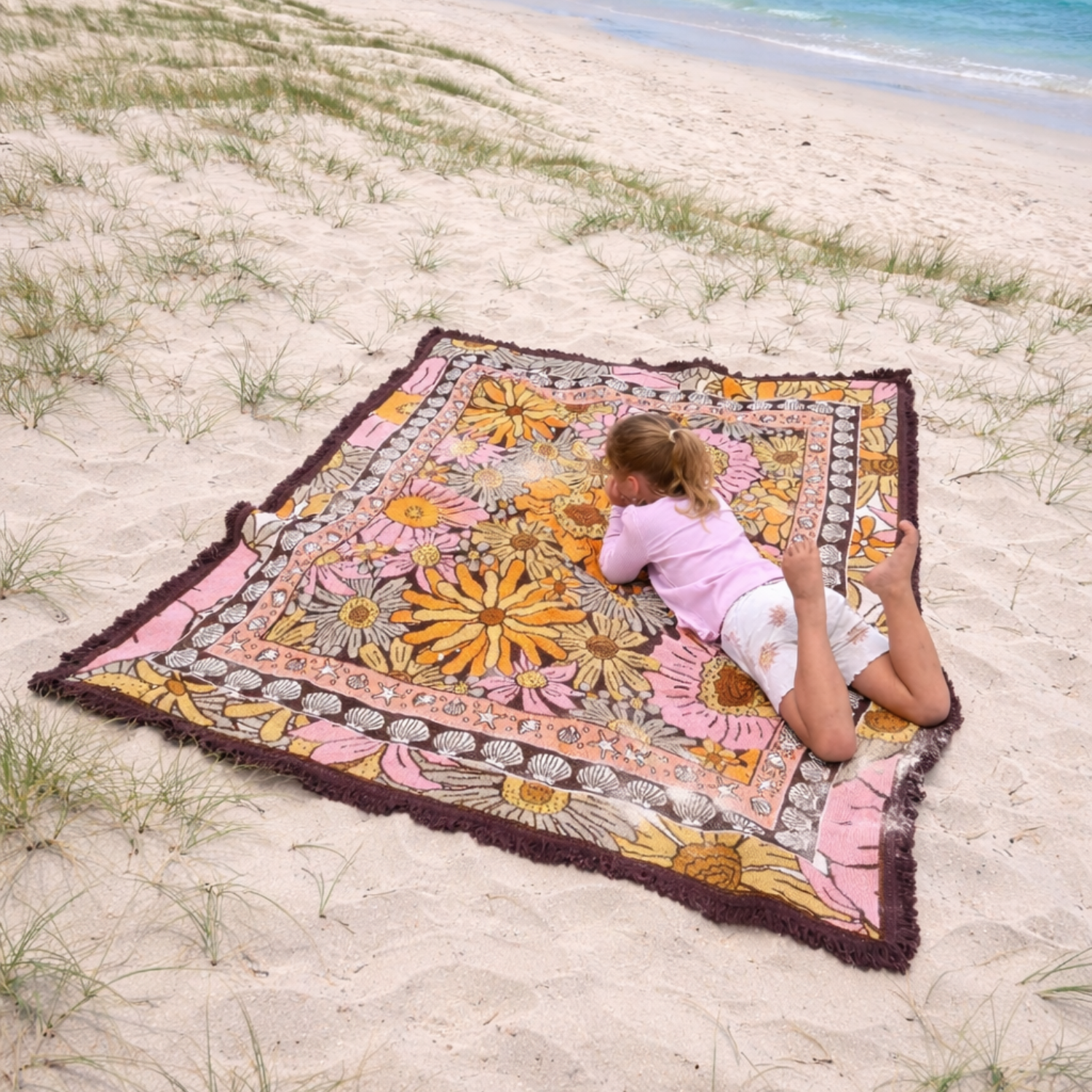 Child relaxing on the Vanaway Sunblossom woven travel throw rug on a sandy beach, enjoying a day by the ocean.