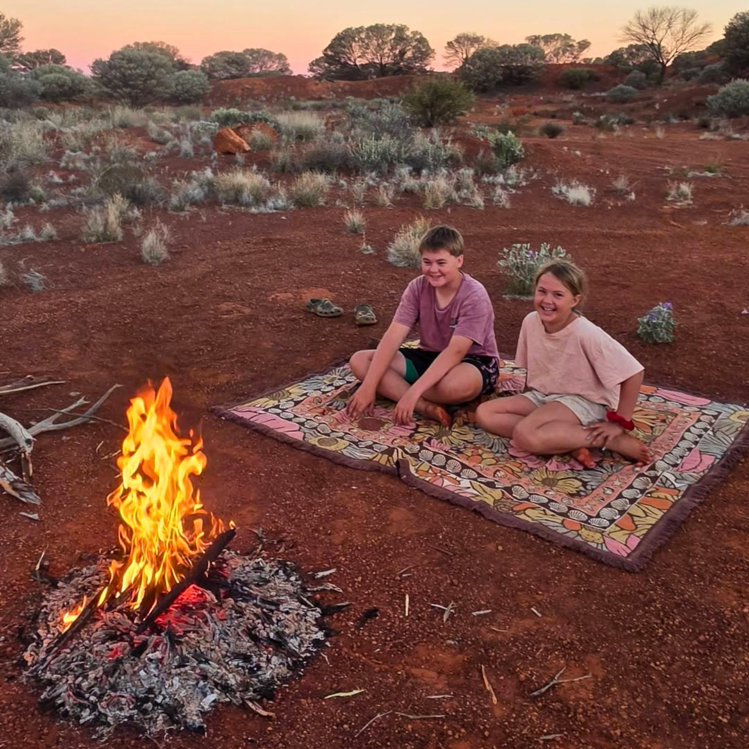 Two children sitting on the Vanaway Sunblossom woven travel throw rug beside a campfire in the Australian outback at sunset.
