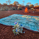 Reverse side of the Vanaway Sunblossom woven travel throw rug laid beside a campfire at an Australian outback campsite, with camping tents in the background at sunset.