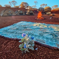 Reverse side of the Vanaway Sunblossom woven travel throw rug laid beside a campfire at an Australian outback campsite, with camping tents in the background at sunset.
