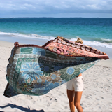 Woman holding the reversible Vanaway Sunblossom woven travel throw rug at the beach, displaying both the vibrant floral front and the reverse design by the ocean.