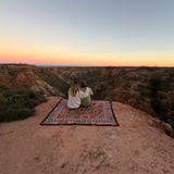 Couple sitting on the Vanaway Sunblossom woven travel throw rug at a scenic cliff lookout, enjoying a sunset over the rugged Australian landscape.