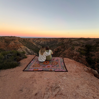 Couple sitting on the Vanaway Sunblossom woven travel throw rug at a scenic cliff lookout, enjoying a sunset over the rugged Australian landscape.