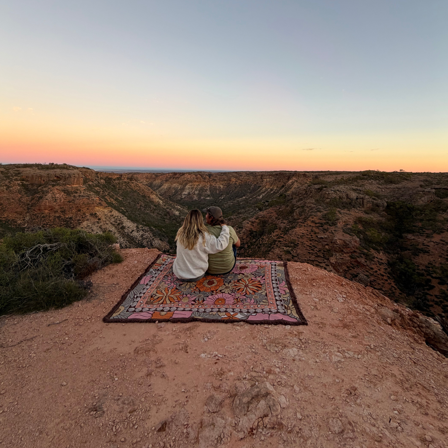 Couple sitting on the Vanaway Sunblossom woven travel throw rug at a scenic cliff lookout, enjoying a sunset over the rugged Australian landscape.