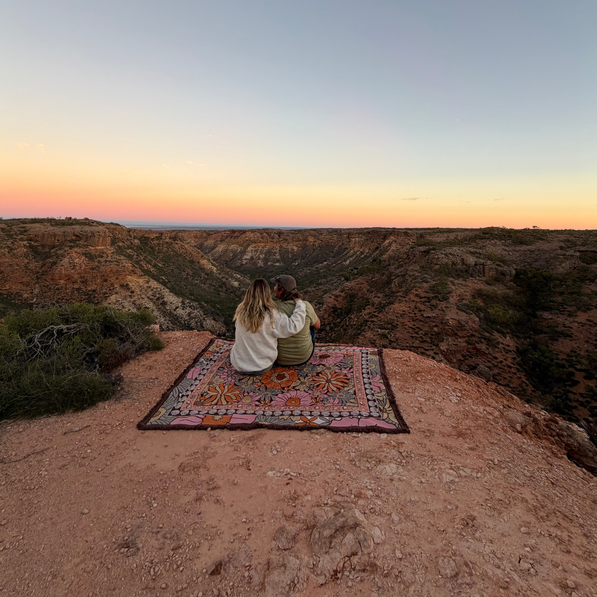 Couple sitting on the Vanaway Sunblossom woven travel throw rug at a scenic cliff lookout, enjoying a sunset over the rugged Australian landscape.
