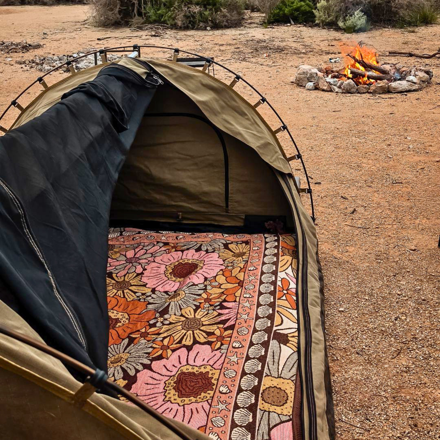 Vanaway Sunblossom woven travel throw rug inside a camping tent with a campfire burning nearby, creating a cosy outdoor camping setup.